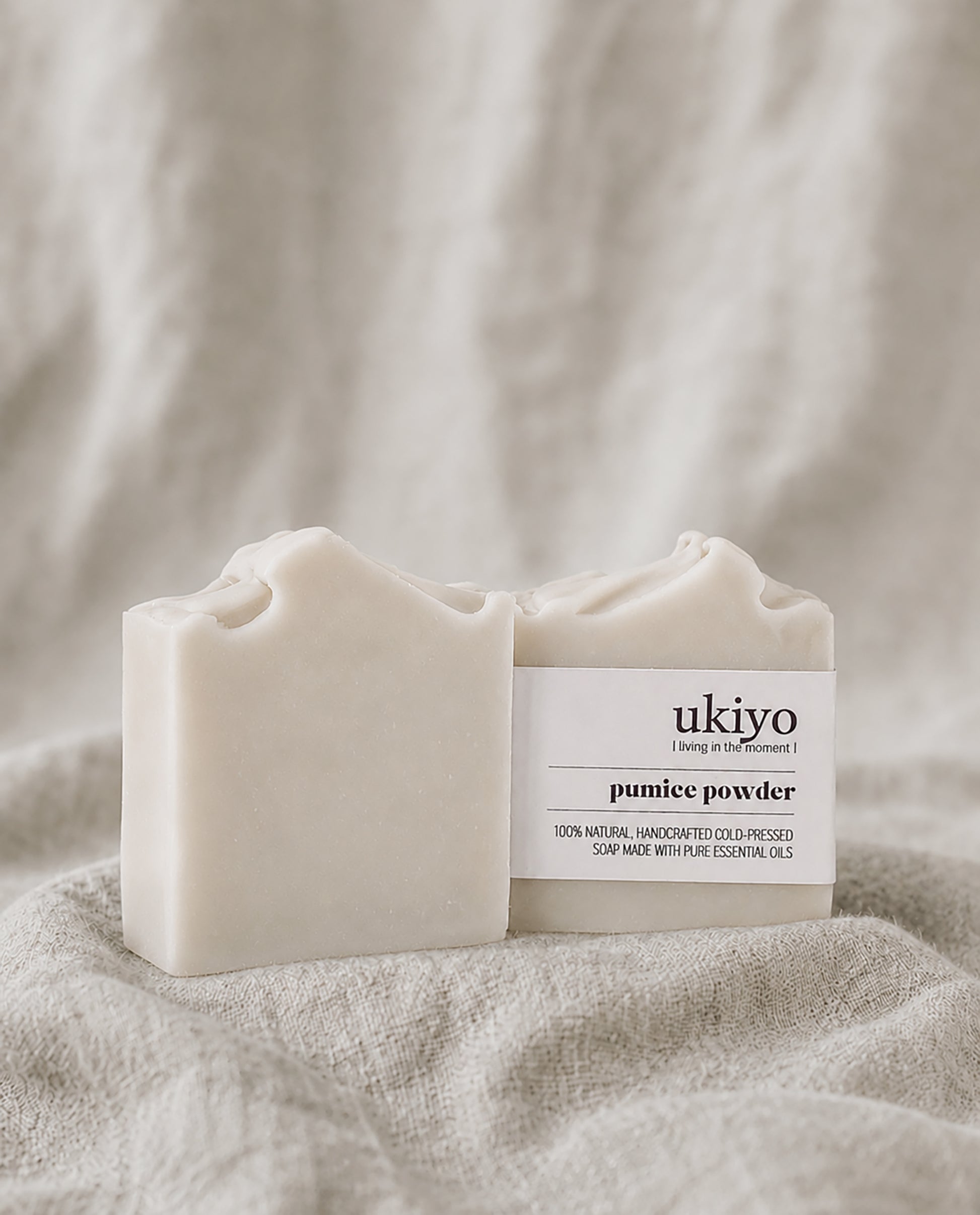 Two bars of pumice powder soap with 'ukiyo' branding on a textured beige fabric background